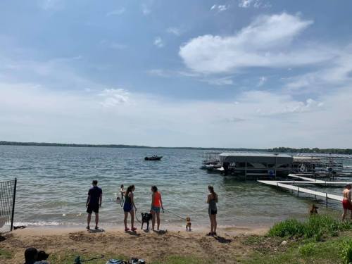 A group of people stands by the shore of a lake, with boats and docks in the background under a partly cloudy sky.