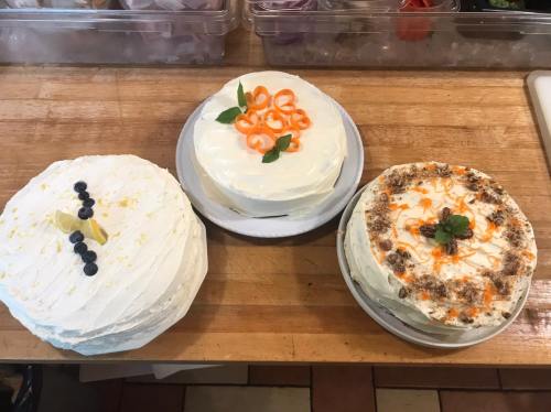 Three decorated cakes on a wooden surface: one with blueberries, one with orange slices, and one with carrot and nuts.