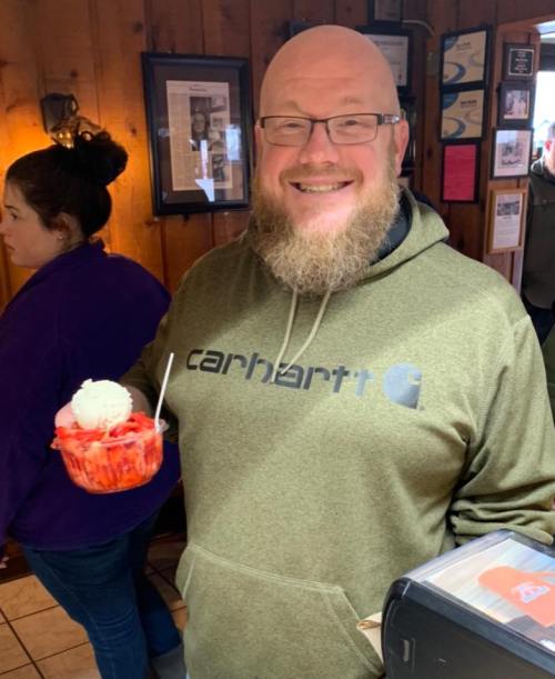 A smiling man with a beard holds a dessert with strawberries and ice cream in a cozy, wooden interior.