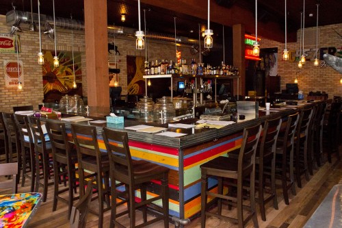 A colorful bar with wooden stools, shelves of liquor, and warm lighting in a cozy restaurant setting.