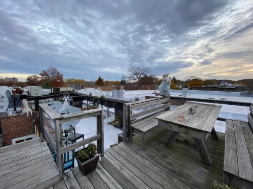 A rooftop view featuring wooden tables, sculptures, and a cloudy sky at sunset.