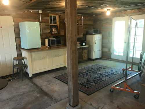 A rustic kitchen with wooden walls, a blue fridge, a white fridge, and a rug on a concrete floor.