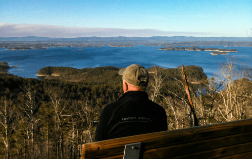 A person sits on a bench overlooking a vast lake surrounded by islands and forested hills under a clear blue sky.