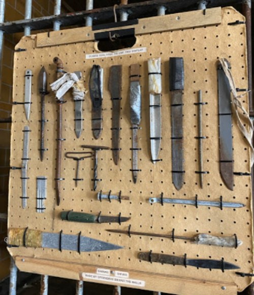 A display board featuring various handmade weapons, including knives and spears, mounted on a wooden background.