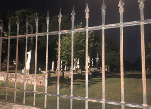 A dark cemetery scene viewed through a rusty iron fence, with gravestones and trees visible in the background.