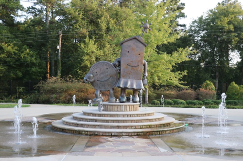 A whimsical statue of a smiling character holding a shield, surrounded by fountains in a park setting.