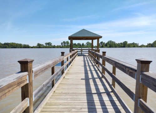 A wooden pier extends over a calm lake, leading to a gazebo under a blue sky with scattered clouds.