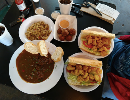 A table with two shrimp po'boys, a bowl of gumbo, rice, and fried balls, accompanied by drinks and utensils.