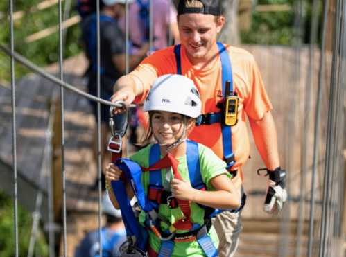 A child in a helmet gives a thumbs up while being guided across a suspension bridge by an adult in safety gear.
