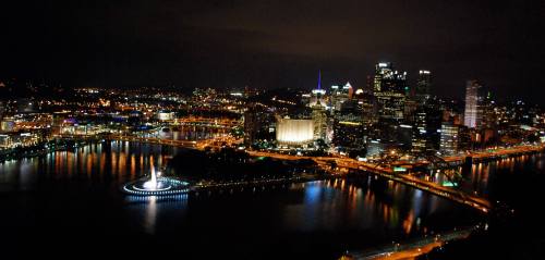 A nighttime view of Pittsburgh's skyline, featuring illuminated buildings and bridges reflecting on the water.