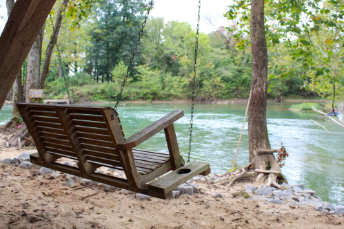 A wooden swing hangs by a river, surrounded by trees and a sandy area, creating a peaceful outdoor scene.