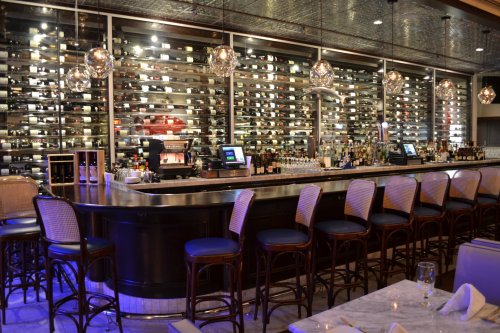A stylish bar with a curved counter, high stools, and a wall of wine bottles illuminated by pendant lights.
