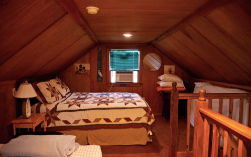 Cozy attic bedroom with wooden walls, a quilted bed, air conditioner, and a small window.