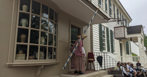 A woman in period clothing stands outside a historic building, holding a pole, with a group of people seated nearby.