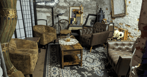 Cozy, vintage-style sitting area with brown chairs, a wooden table, and decorative items against a rustic wall.