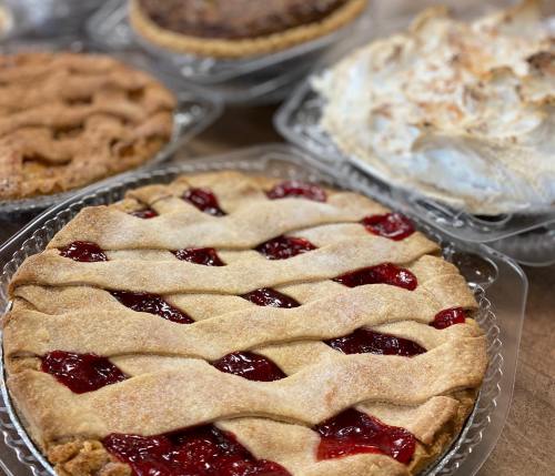 A close-up of a cherry pie with a lattice crust, surrounded by other assorted pies on a table.