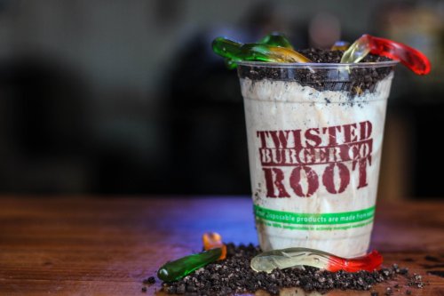 A cup of root beer float topped with crushed cookies and gummy worms, placed on a wooden surface.