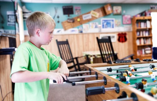 A young boy plays foosball in a cozy room filled with books and wooden furniture.