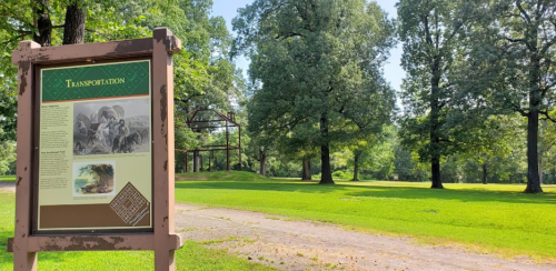 A sign about transportation stands in a green park with trees and a path, under a clear blue sky.