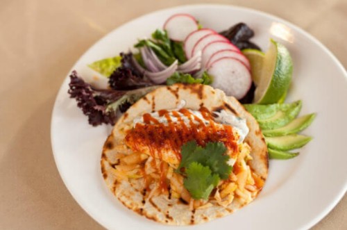 A plate featuring a taco with shredded chicken, topped with sauce and cilantro, alongside fresh vegetables and lime wedges.