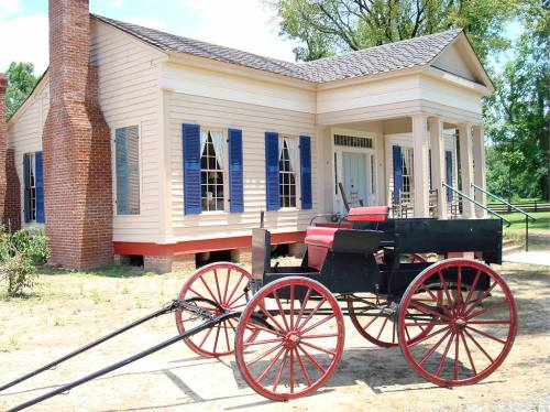 A historic building with blue shutters and a red and black horse-drawn carriage in front on a sunny day.