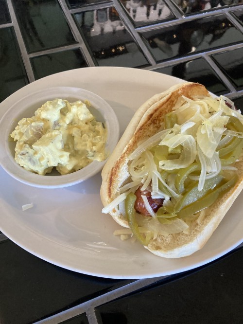 A hot dog in a bun topped with onions and green peppers, served with a side of creamy potato salad.
