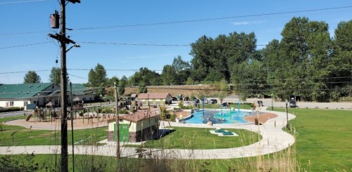 A sunny park featuring a splash pad, playground, and green spaces, surrounded by trees and buildings.