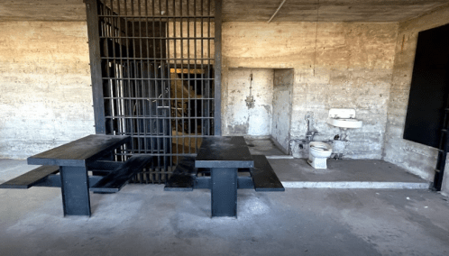 Interior of a prison cell with a barred door, two tables, a toilet, and a sink against a concrete wall.