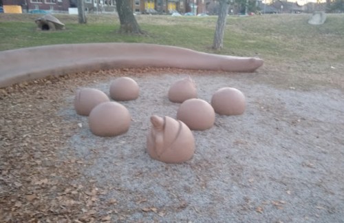 A playground with several rounded, clay-like sculptures on a gravel surface, surrounded by grass and trees.