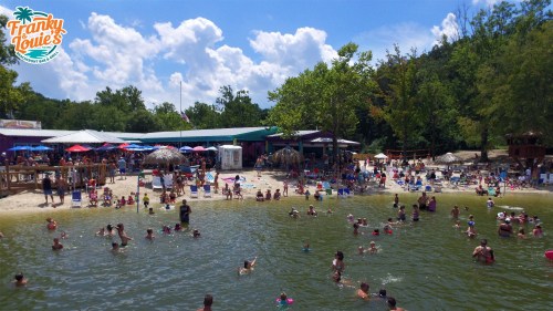 A busy beach scene with people swimming, lounging, and enjoying the sun, surrounded by trees and colorful umbrellas.