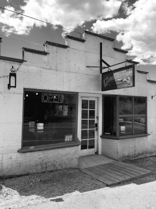 A black and white photo of a small, open storefront with a sign and large windows on a sunny day.