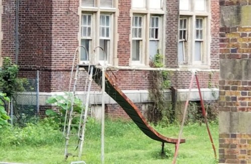 Abandoned playground with a rusty slide and swing set, overgrown grass, near an old brick building.