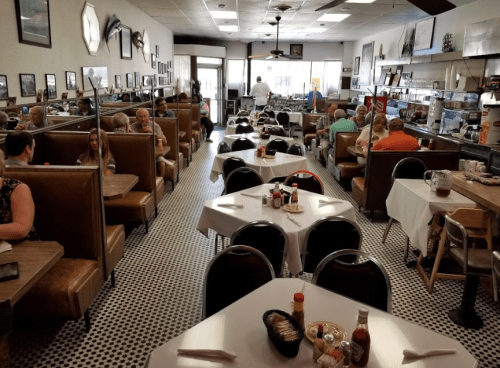 A busy diner with booths and tables, featuring patrons enjoying meals and a staff member serving in the background.