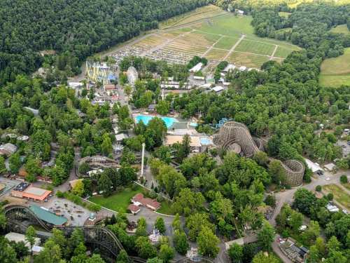 Aerial view of a theme park surrounded by lush greenery, featuring rides, a pool, and open fields.