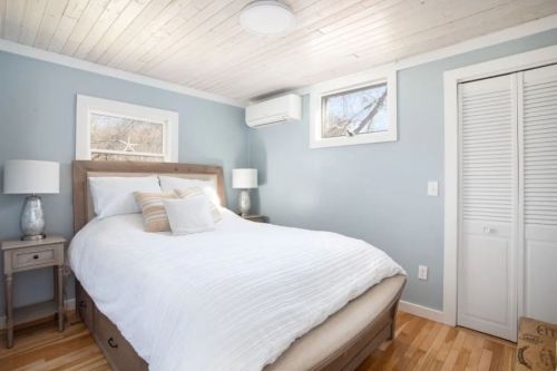 Cozy bedroom with a queen bed, light blue walls, wooden floors, and two windows letting in natural light.