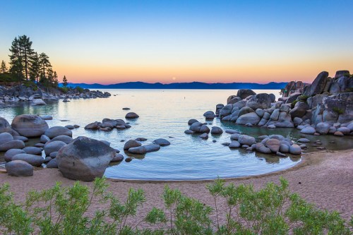 Serene lakeside view at sunset, featuring smooth rocks, calm water, and a colorful sky over the horizon.