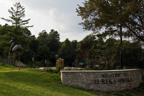 A stone welcome sign for Eureka Springs, surrounded by trees and greenery, with a large bird sculpture nearby.