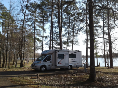 A parked RV surrounded by trees near a calm lake on a sunny day.