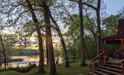 A serene riverside scene at sunset, featuring trees, a cozy cabin, and a hammock. Soft lights illuminate the porch.
