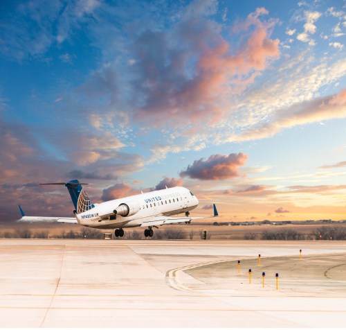 A United Airlines plane takes off against a colorful sunset sky, with clouds and a clear runway in the foreground.
