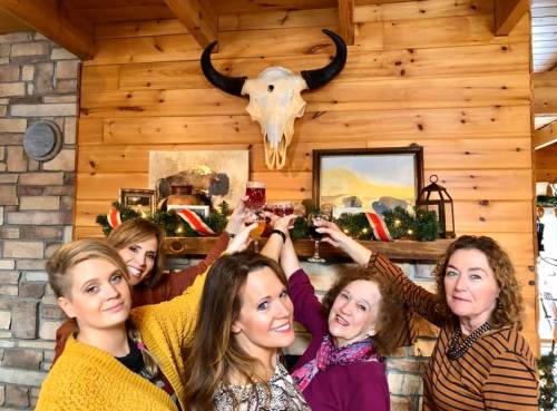 Five women toast with drinks in a cozy, rustic setting, featuring a bull skull and festive decorations on the wall.
