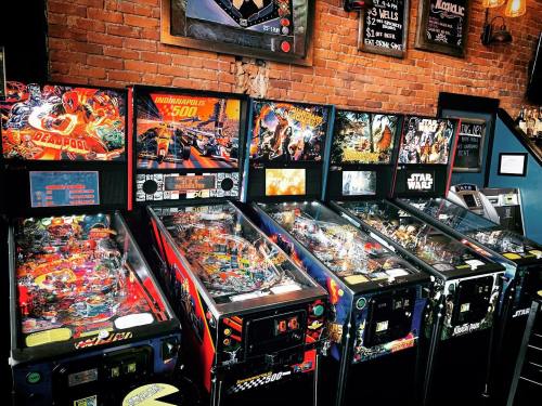A row of colorful pinball machines featuring themes like Deadpool and Star Wars, set against a brick wall.