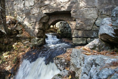 A stone bridge arching over a flowing stream, surrounded by rocks and autumn leaves.
