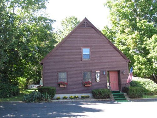 A brown, two-story house with a steep roof, green steps, and flower boxes, surrounded by trees and an American flag.