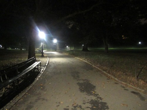A dimly lit park path at night, lined with benches and illuminated by street lamps, surrounded by trees.