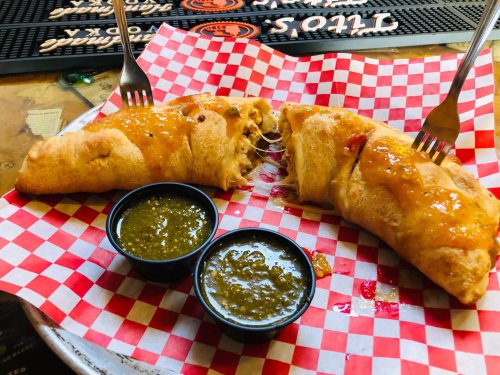 A plate with two cheesy calzones drizzled with sauce, served with two small cups of green sauce on a checkered paper.
