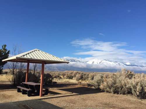 A picnic shelter with a metal roof overlooks a snowy mountain range under a clear blue sky. Sagebrush surrounds the area.