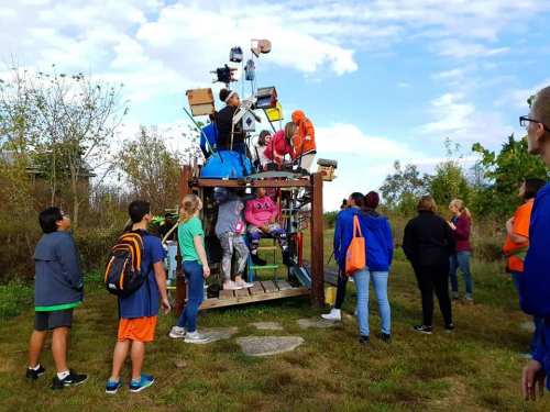 A group of people gathers around a large, whimsical sculpture made of various objects in a grassy outdoor setting.
