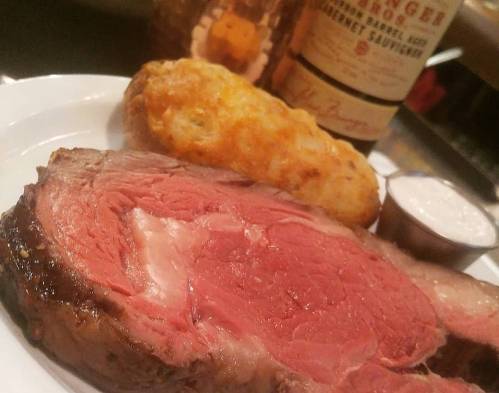 A plate featuring a slice of juicy prime rib, a crispy potato dish, and a small cup of dipping sauce.