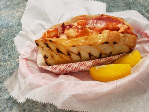 A grilled lobster roll served in a toasted bun, accompanied by two lemon wedges on a paper-lined tray.